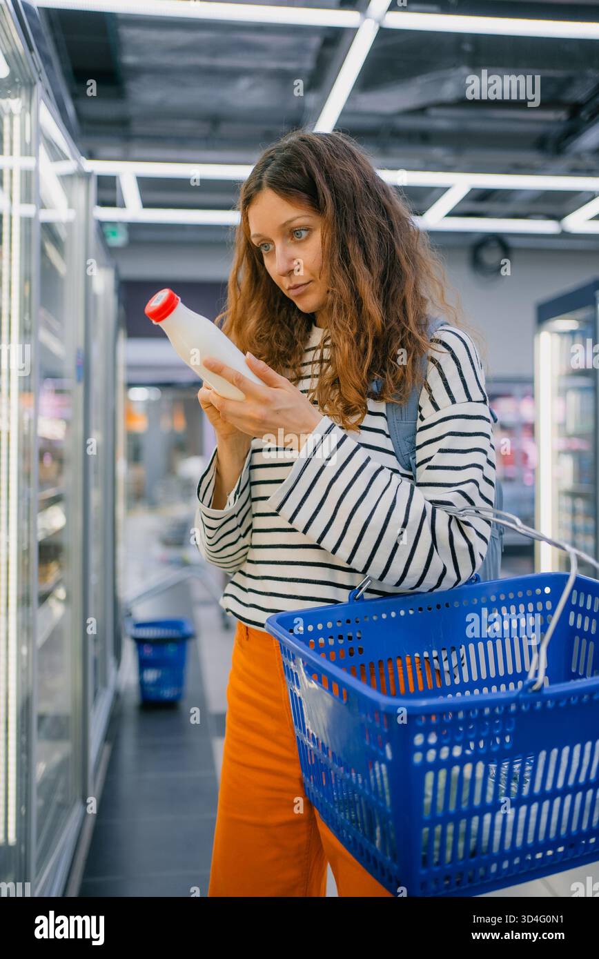 Donna in piedi in un corridoio di un supermercato, concentrandosi intensamente sull'etichetta di un cartone di latte, tenendo in mano un cestino blu mentre prende decisioni informate sui suoi acquisti di generi alimentari Foto Stock