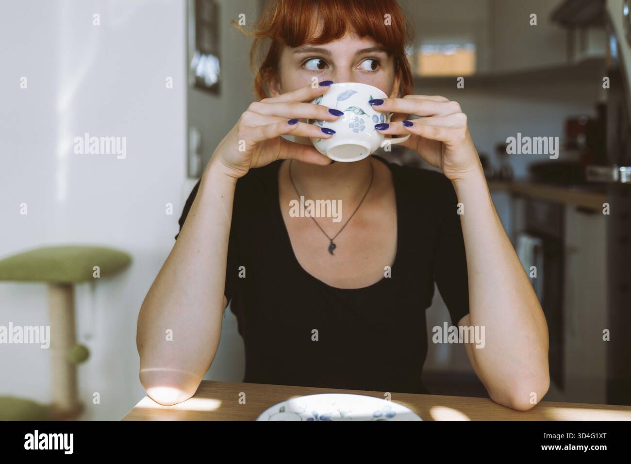Ritratto giovane donna con i capelli rossi legati in panino seduto a tavola, tenendo la tazza e il piattino in mano, bevendo tè verde, infusione di erbe, caffè, mattina Foto Stock