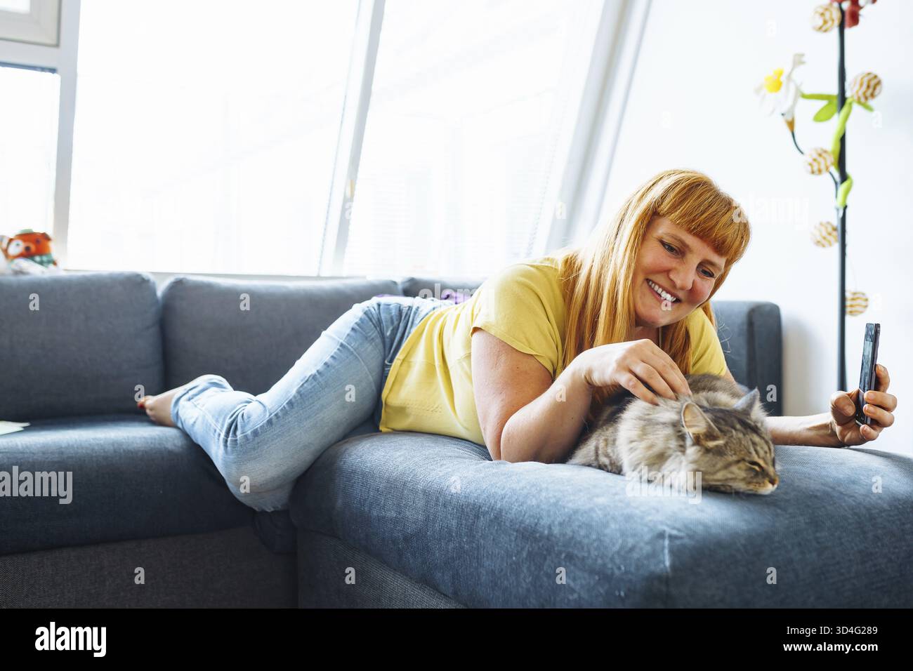 Ritratti una donna bionda di mezza età sdraiata sul divano abbracciando un gatto che scatta selfie. la vita a casa con un animale domestico Foto Stock
