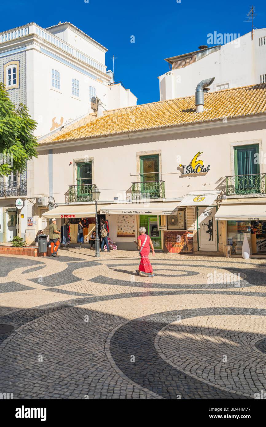 Lagos, Portogallo, 8 novembre 2025, Una donna in rosso attraversa una piazza illuminata dal sole con ciottoli bianchi e neri a onde nel centro della città. donna io Foto Stock