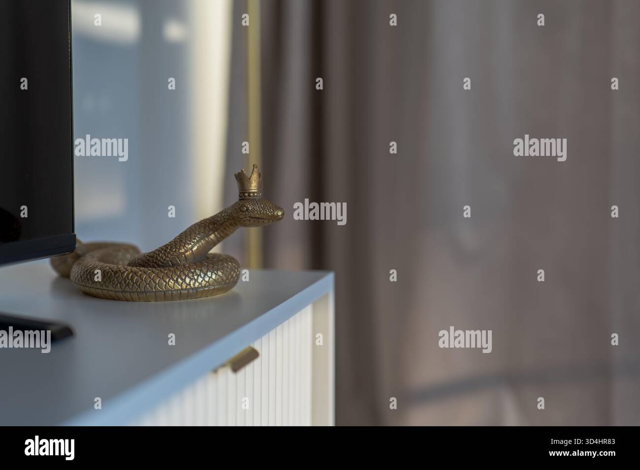 Primo piano mostra un serpente sormontato da una corona d'oro su un armadio blu pallido a Danzica. La luce soffusa della sera attraverso le tende trasparenti proietta ombre calde in un design minimale Foto Stock