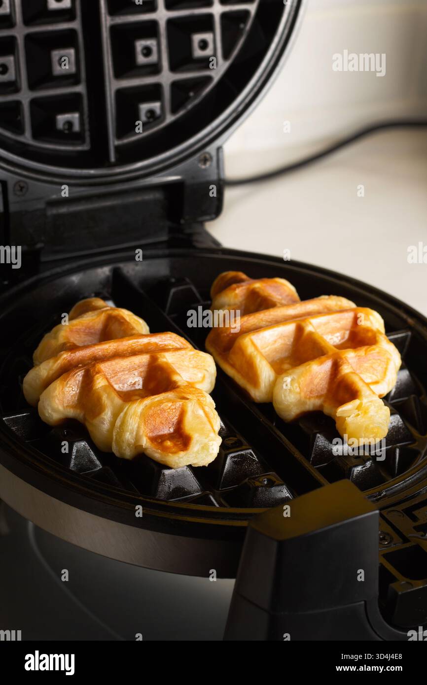 Due waffle dorati cucinano in una macchina per waffle neri sul piano della cucina. La cucina calda e invitante suggerisce una deliziosa esperienza di colazione Foto Stock