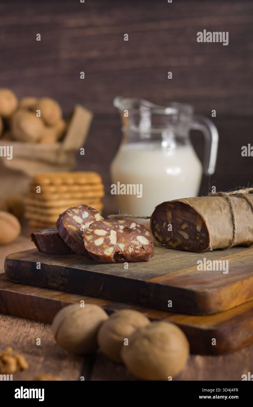 I rotoli di cioccolato e noci affettati riposano su una tavola di legno accanto a una caraffa di latte e a una pila di biscotti, creando un ambiente caldo e invitante per uno spuntino. Foto Stock