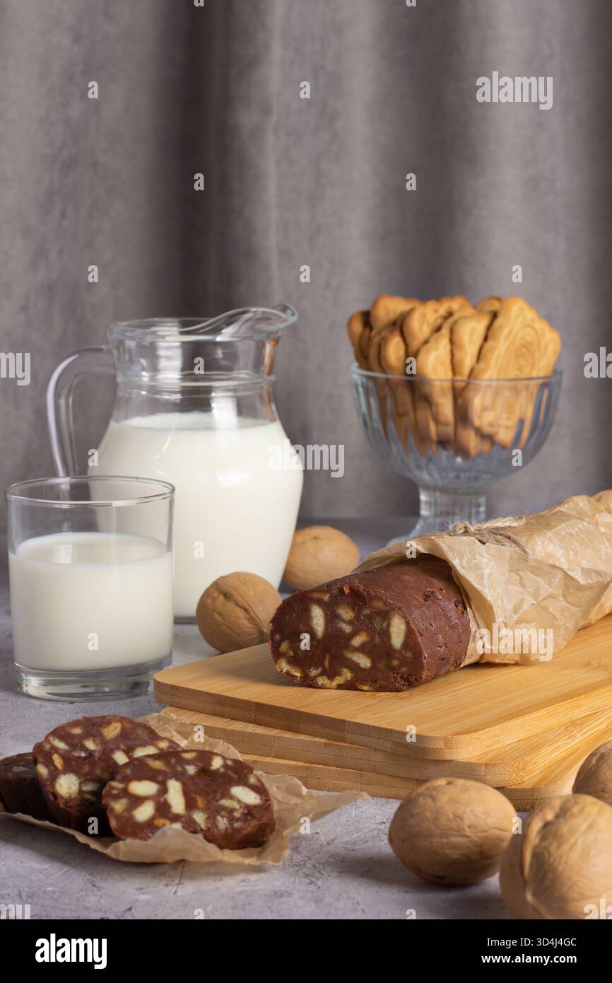 Il salame al cioccolato viene tagliato a fette e posto su un asse di legno accanto a un bicchiere di latte. Una caraffa di latte e una ciotola di biscotti sono dolci e invitanti Foto Stock