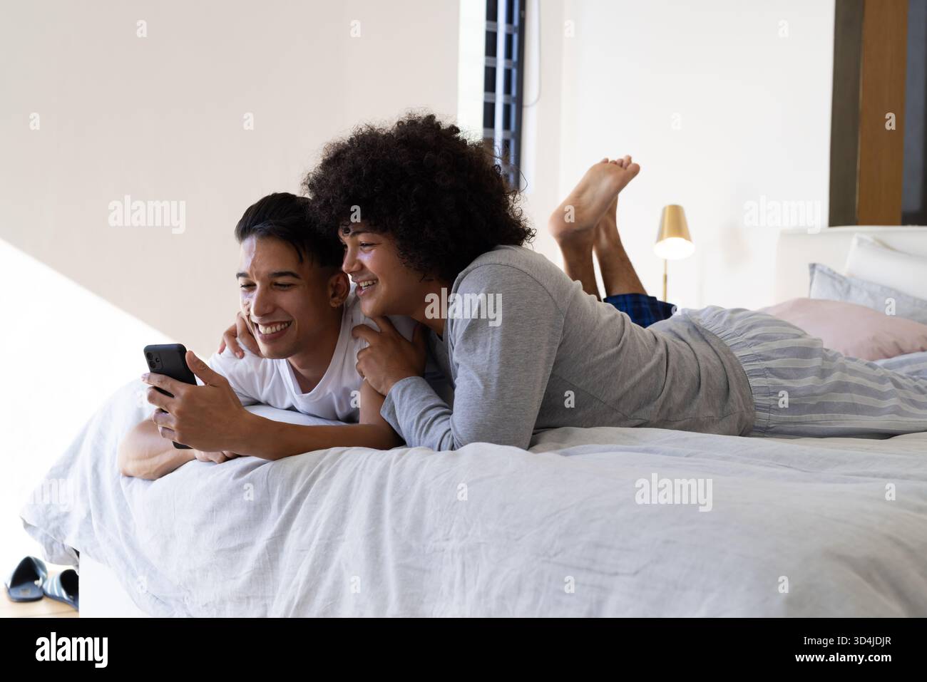 Sdraiati diversi partner maschili che si coccolano sul letto di casa tenendo in mano lo smartphone con cuscini e lampada color pastello Foto Stock