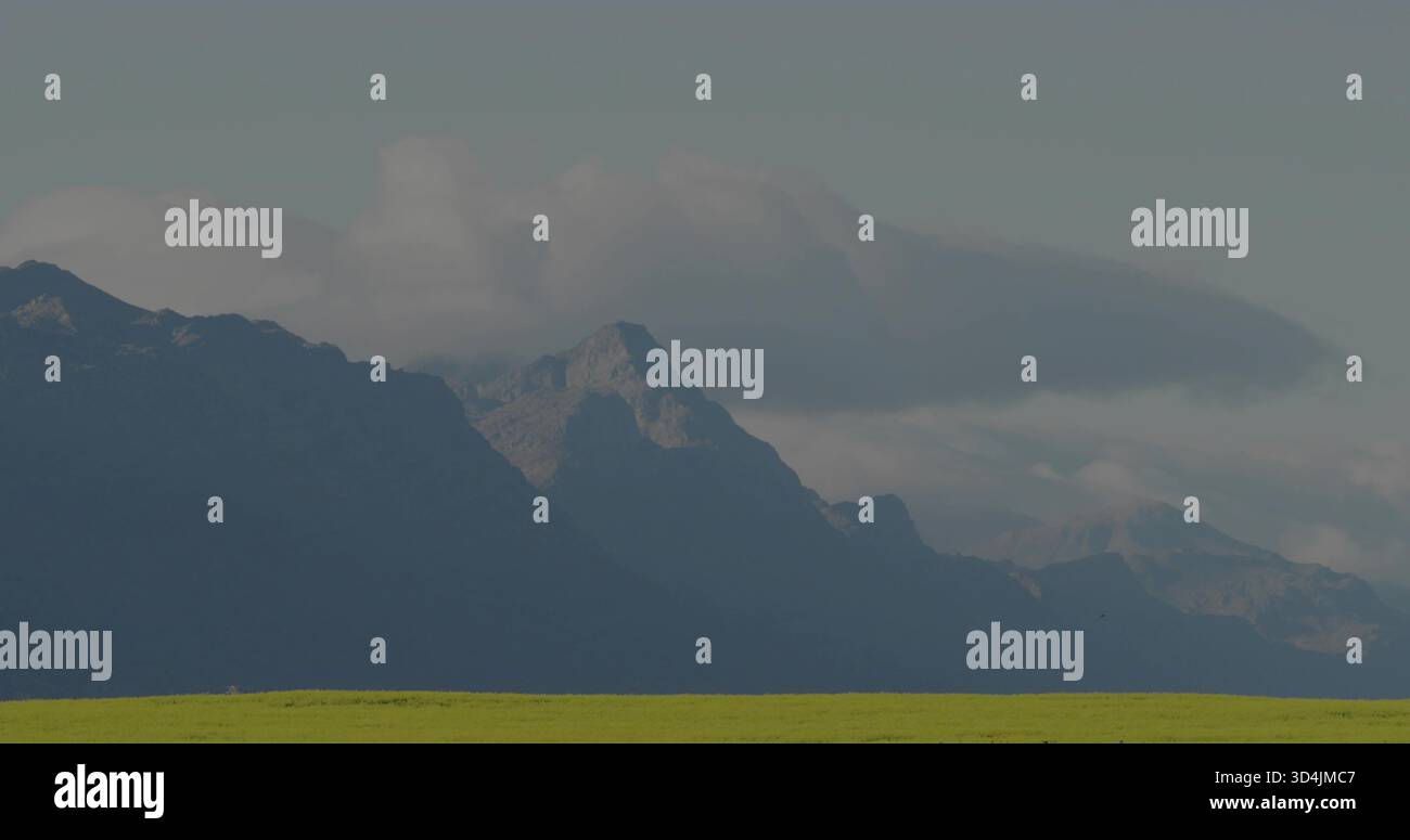 Mostra una catena montuosa a strati che si innalza sopra un prato erboso ai piedi delle colline, con basse nuvole e nebbia Foto Stock