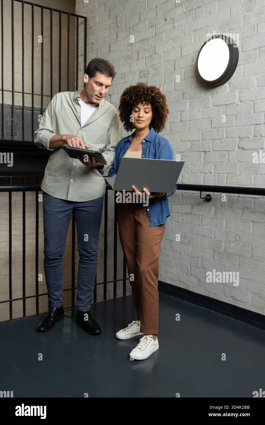 Diversi colleghi che esaminano i contenuti digitali sull'atterraggio delle scale, con notebook e tablet Foto Stock