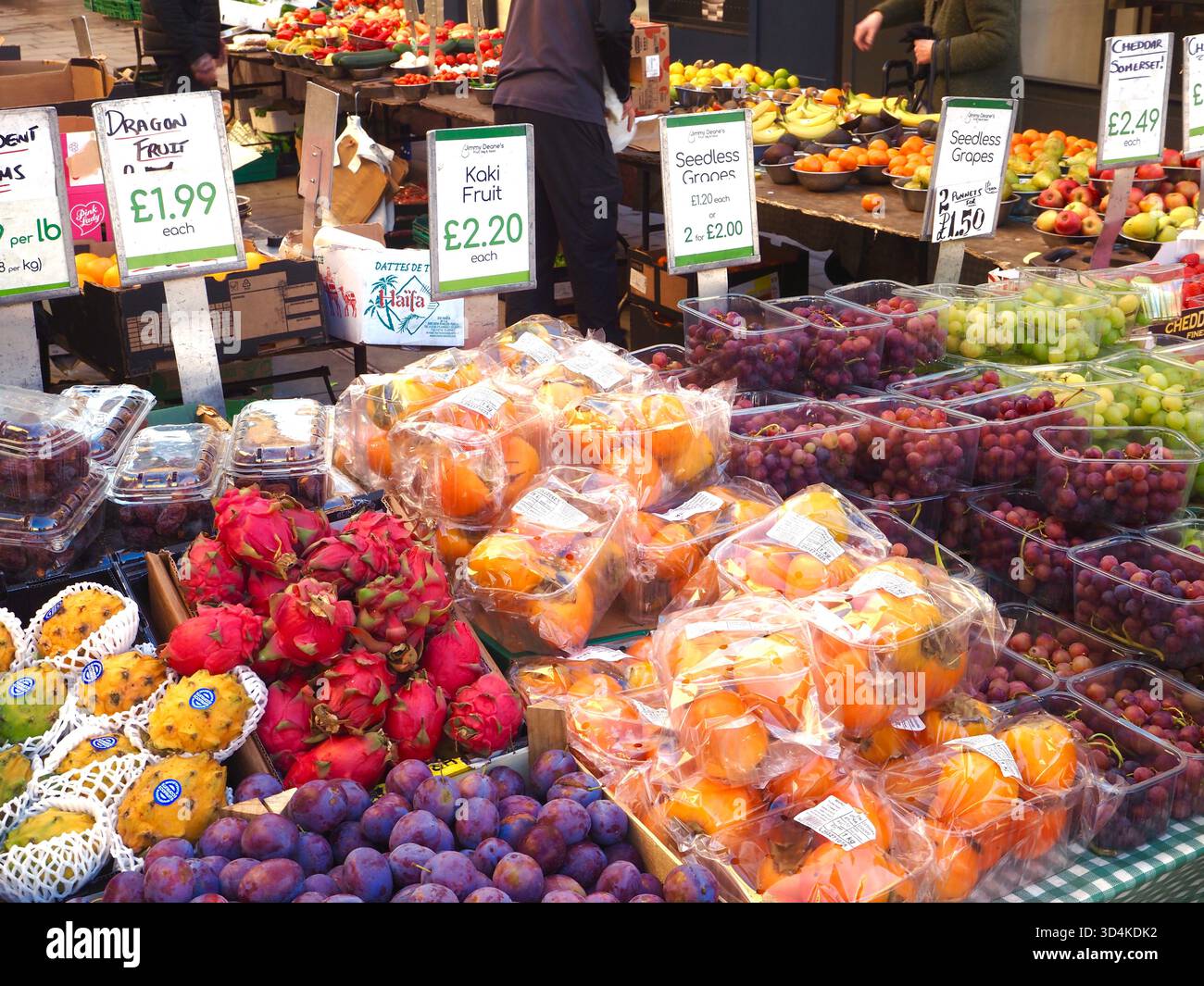 Frutta dai colori vivaci in vendita su una bancarella di frutta e verdura con etichette dei prezzi scritte a mano e clienti e venditori sullo sfondo Foto Stock