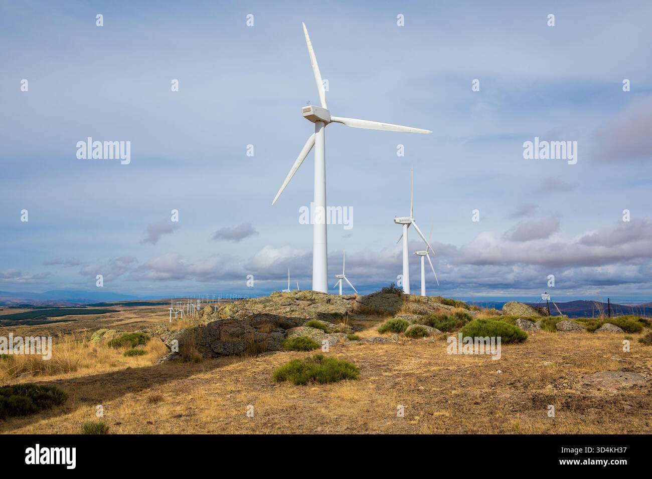 Diverse turbine eoliche si innalzano su un paesaggio erboso con affioramenti rocciosi, mostrando una produzione energetica sostenibile sotto un cielo luminoso e parzialmente nuvoloso Foto Stock