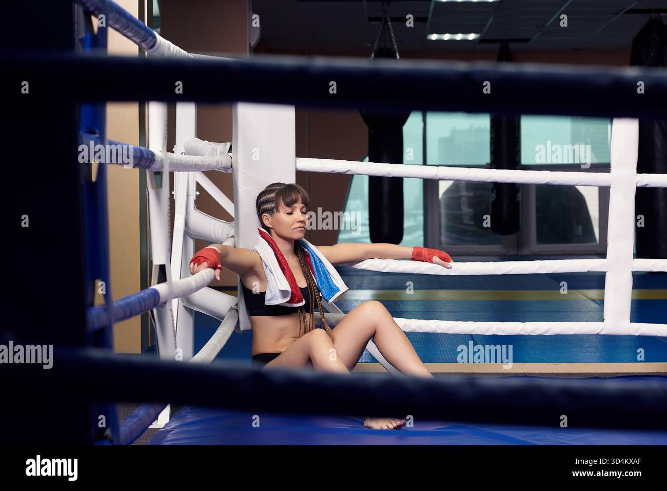 Una lottatrice con la benda da boxe sulle mani siede in un anello di boxe. La donna ha un asciugamano sul collo. Foto Stock