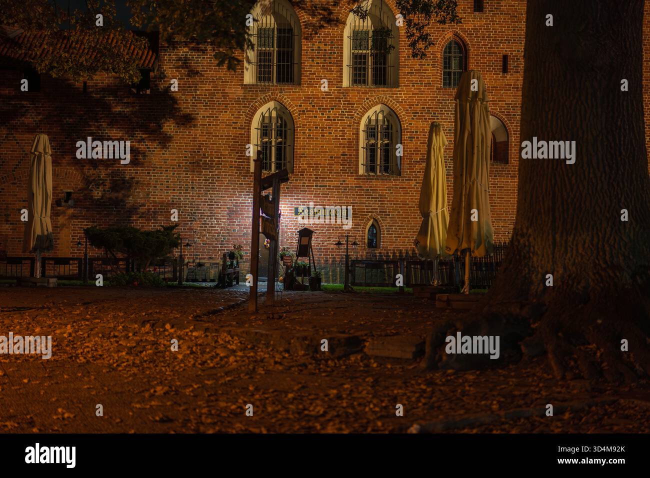 Vista notturna del castello di Malbork a Malbork, Polonia, luci calde su mattoni rossi, finestre ad arco, insegna Restauracja, foglie cadute, ombrelli chiusi, alberi, s. Foto Stock
