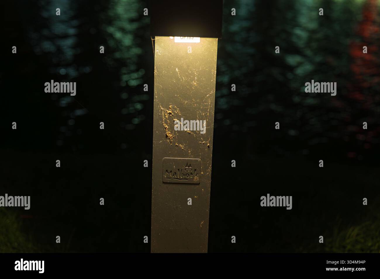 Una luce a dissuasore ravvicinata illumina un palo metallico sottile di notte vicino all'acqua vicino al fiume Malbork Nogat, un caldo bagliore sui graffi, le increspature riflettono verde e blu Foto Stock