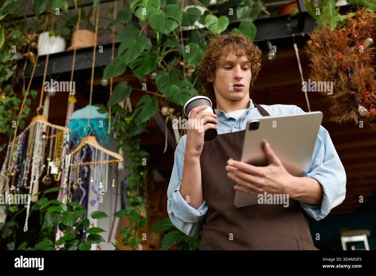 Il giovane in grembiule usa un tablet mentre tiene in mano una tazza di caffè in un ambiente vivace e creativo Foto Stock