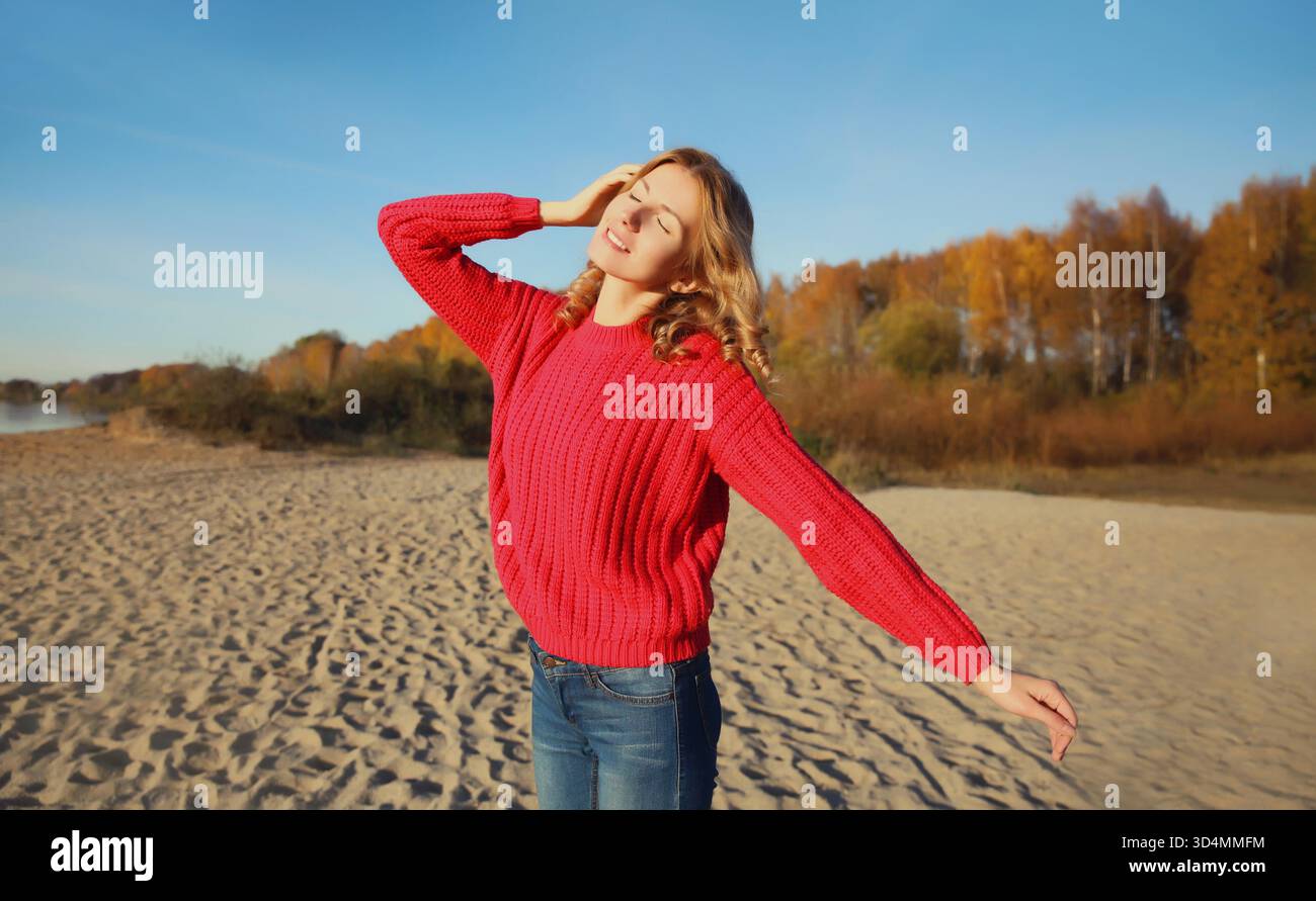 Felice sorridente giovane donna che cammina sulla spiaggia, gode del caldo sole, indossa un maglione in piedi sulla costa del mare, contro alberi con foglie gialle, sfondo naturale || modello rilasciato Foto Stock