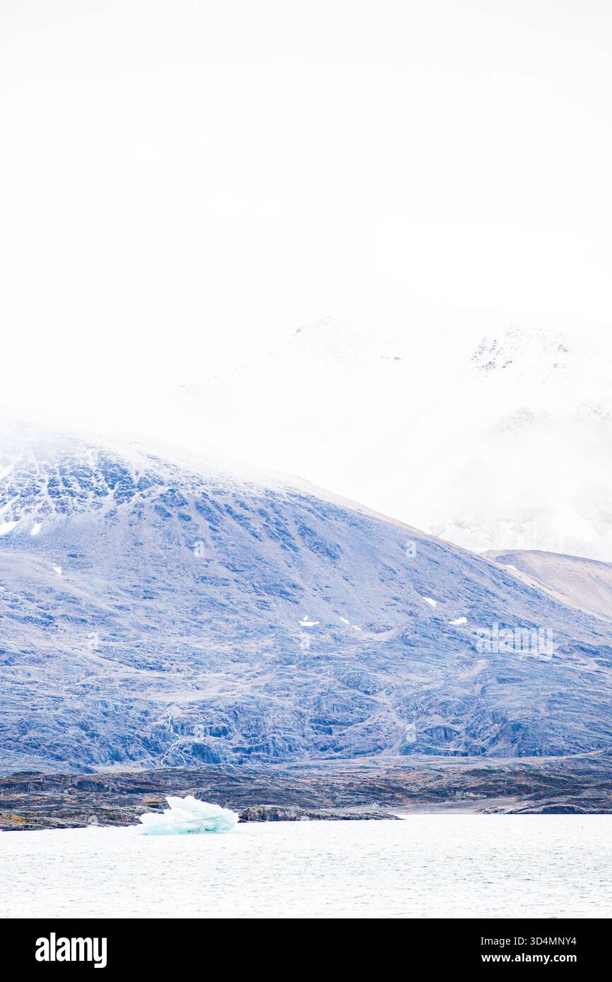 Crociera alle Svalbard - catena montuosa con un cielo blu e un grande blocco di ghiaccio che galleggia nell'acqua. La scena è serena e pacifica, con le montagne che lo circondano Foto Stock
