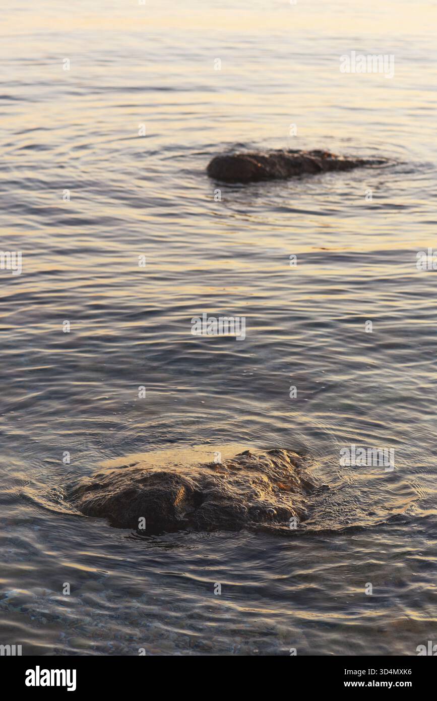 Rocce costiere sommerse in acque tranquille al tramonto, catturando il movimento sereno e la calda luce serale sulla superficie. Messa a fuoco selettiva. Foto Stock