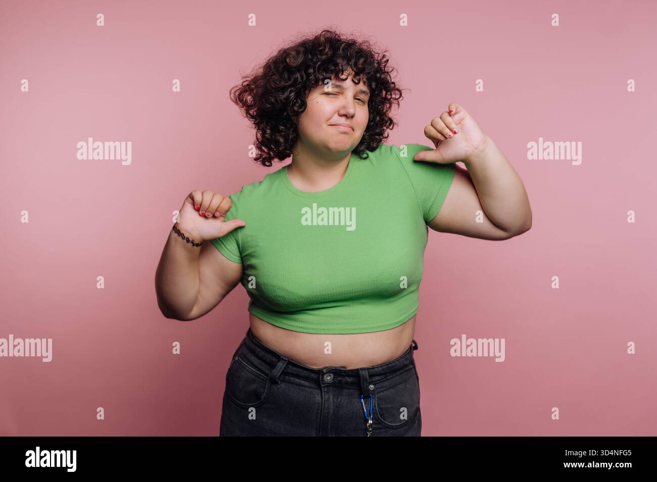 Una donna con i capelli ricci si trova davanti a uno sfondo rosa. Indossa una camicia verde e mostra un gesto verso il basso con i pollici, esprimendo disprezzo o disapprovazione Foto Stock