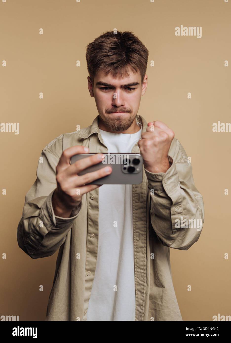 Un giovane con i capelli corti mostra entusiasmo stringendo il pugno mentre guarda un evento sportivo sul suo smartphone. Lo sfondo è un semplice beige co Foto Stock