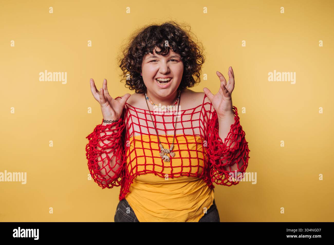 Una persona con capelli ricci posa gioiosamente in una creativa maglia rossa sopra una camicia gialla. Lo sfondo giallo brillante esalta l'atmosfera giocosa. Foto Stock