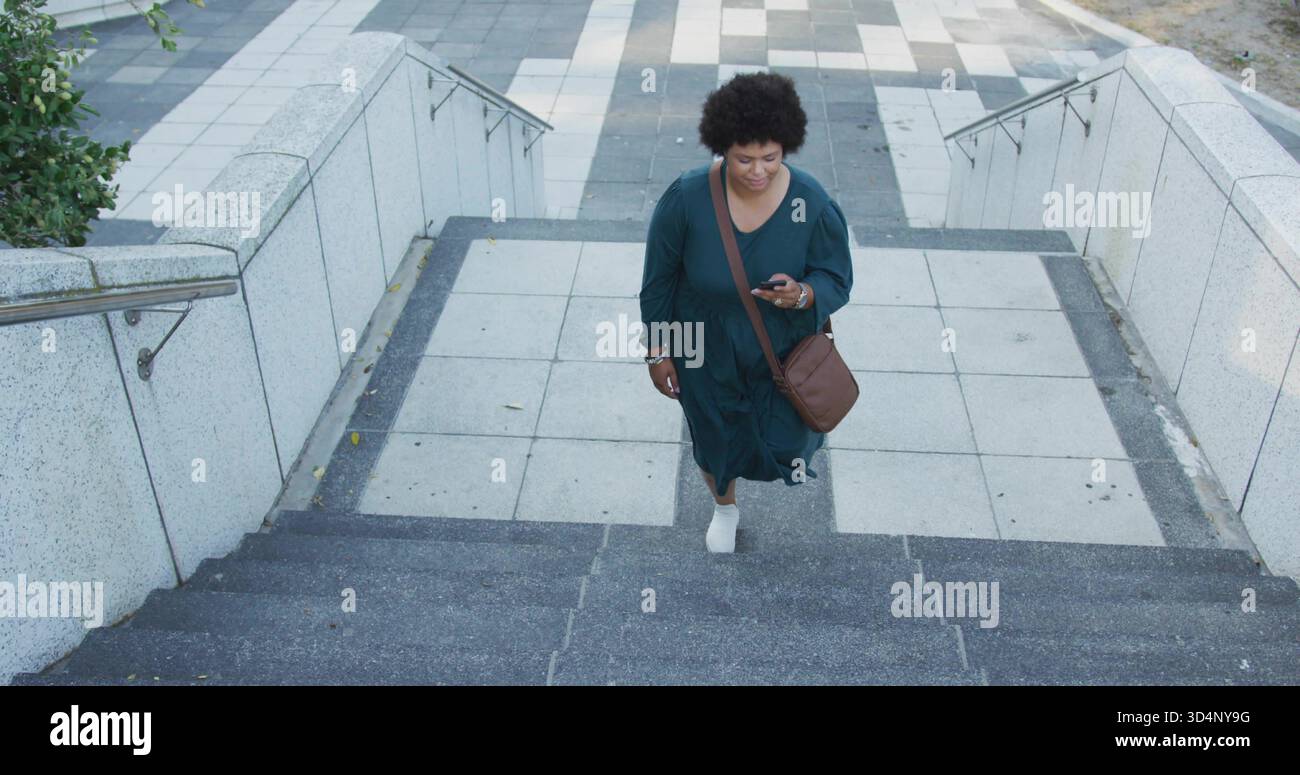 Donna arrampicata in un abito color ottanio che sale gradini di cemento fino alla piazza piastrellata, tenendo in mano il telefono e la borsa marrone Foto Stock