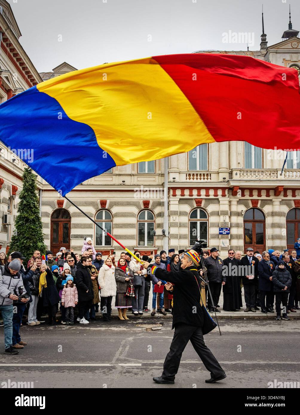 Uomo che sventola bandiera rumena durante la parata nazionale di Bacău, circondato dalla folla e dagli edifici storici. Foto Stock