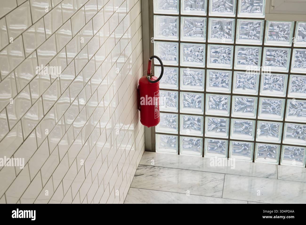 Estintore rosso su pareti piastrellate in interni moderni con blocchi di vetro e pavimento in marmo, equipaggiamento di sicurezza minimo in corridoio luminoso Foto Stock