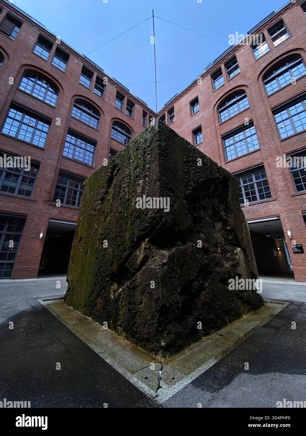 Scultura in pietra ricoperta di muschio in un cortile incorniciato da edifici in mattoni rossi con finestre ad arco. - Immagine stock catturata con smartphone