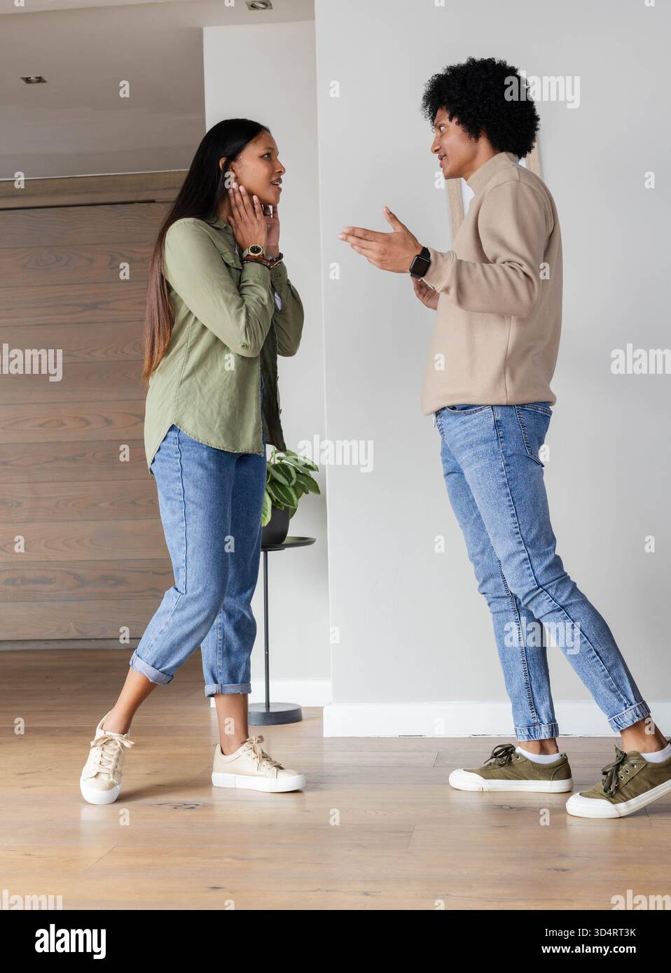 Parla con amici diversi in piedi sul pavimento in legno di casa, con porta scorrevole, piante e smartwatch Foto Stock