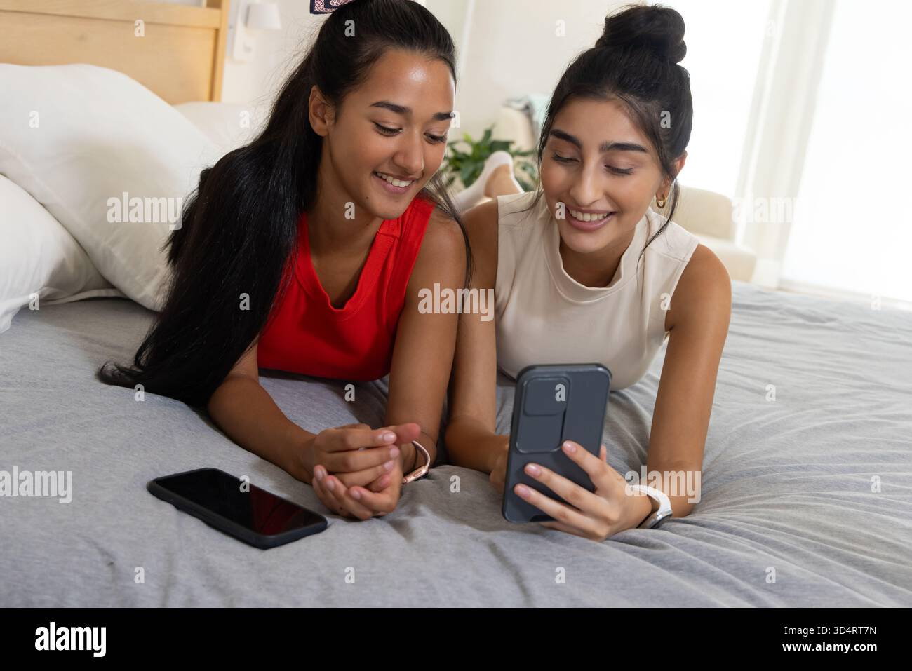 Sdraiate diverse amiche che usano lo smartphone a letto in una camera luminosa, con il telefono di riserva Foto Stock