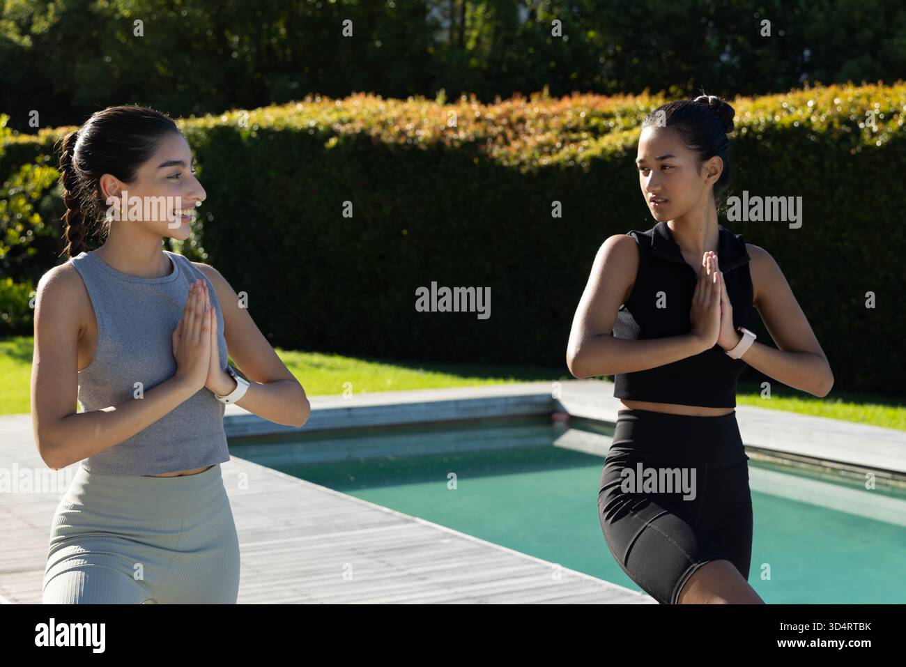 Diverse amiche che praticano la posa sull'albero sul ponte della piscina nel cortile posteriore, con smartwatch Foto Stock