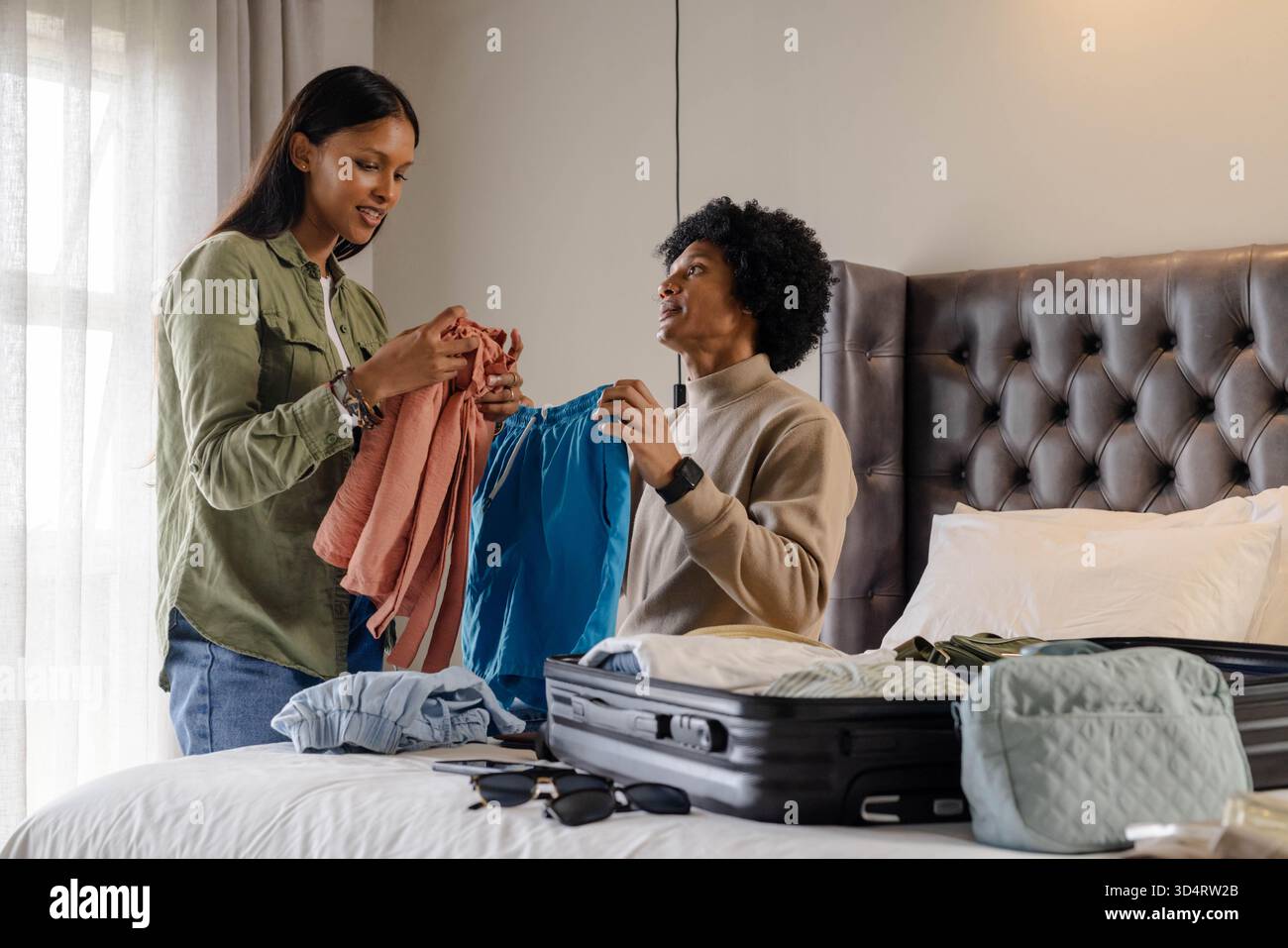 Imballare diverse coppie smistando vestiti sul letto in camera da letto, con valigia nera aperta e camicetta rosa Foto Stock