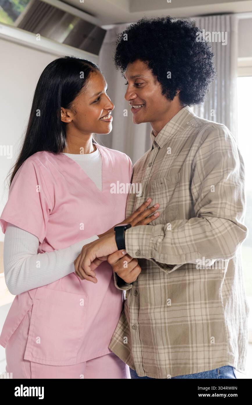 Sorridenti coppie diverse che condividono i momenti nel moderno soggiorno, con smartwatch nero e scrub rosa Foto Stock