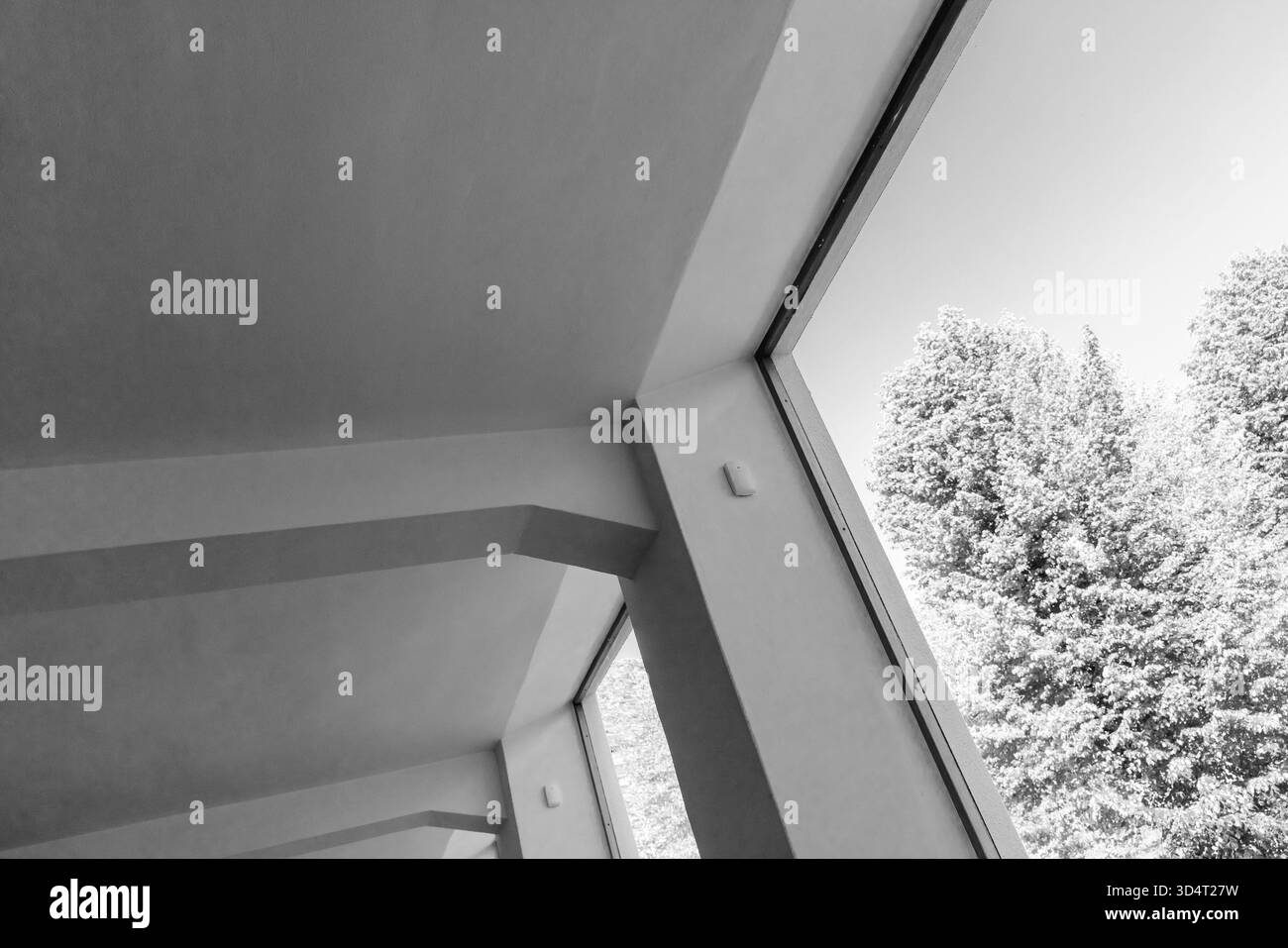 Un ambiente interno bianco e nero caratterizzato da pareti angolate e ampie finestre che incorniciano alberi frondosi all'esterno, creando linee pulite, luce naturale, e. Foto Stock