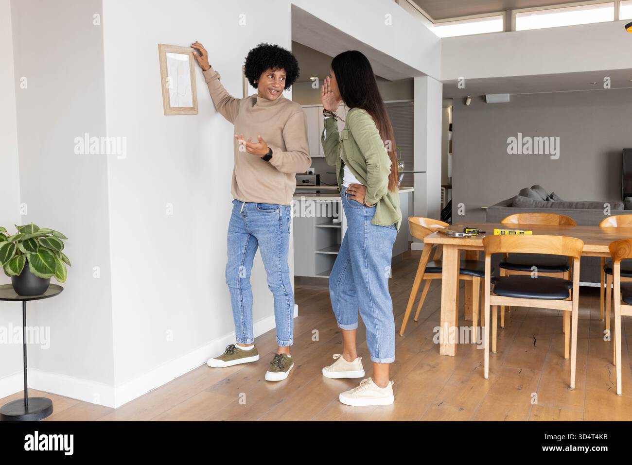 Diverse amiche che discutono di posizionamento delle immagini in una casa a pianta aperta, con cornice e livello in legno Foto Stock