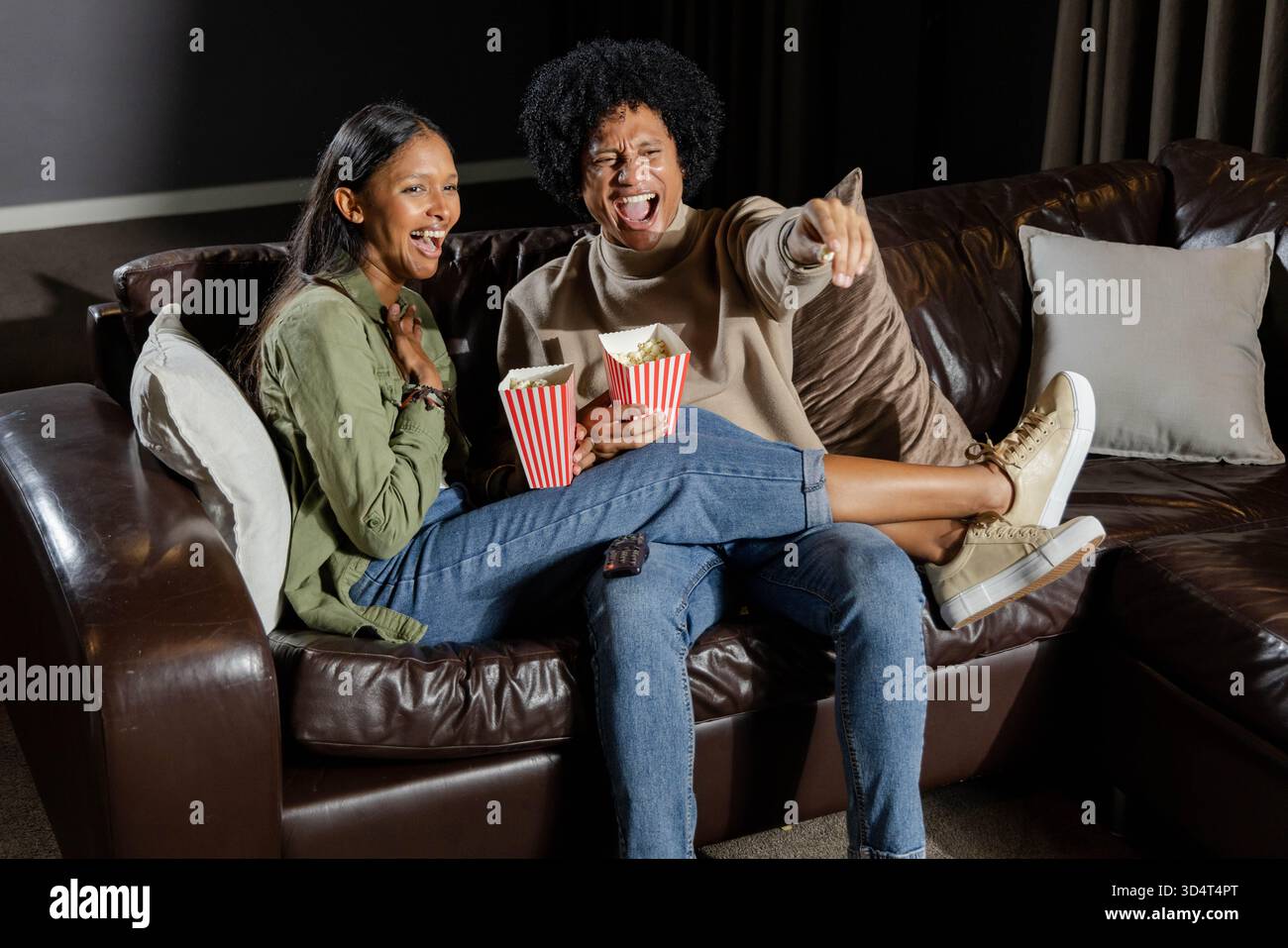 Una coppia di persone che si diverte a ridere sedute su un divano in pelle marrone scuro nel sistema Home Theater, tenendo in mano scatole di popcorn Foto Stock
