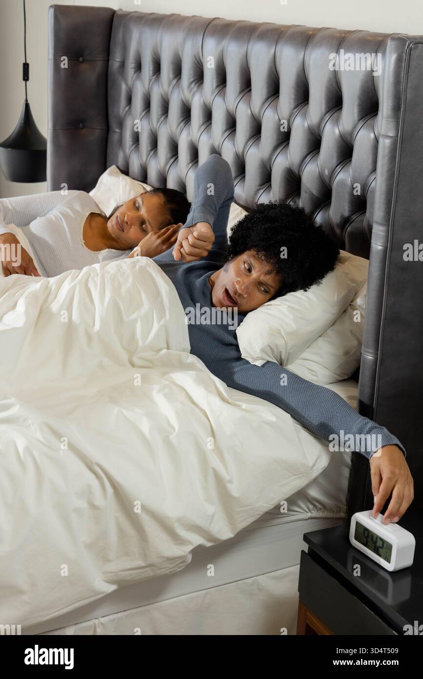Sbadiglianti coppie diverse sdraiate su un letto king size contro la testata grigia, che si avvicinano alla sveglia Foto Stock