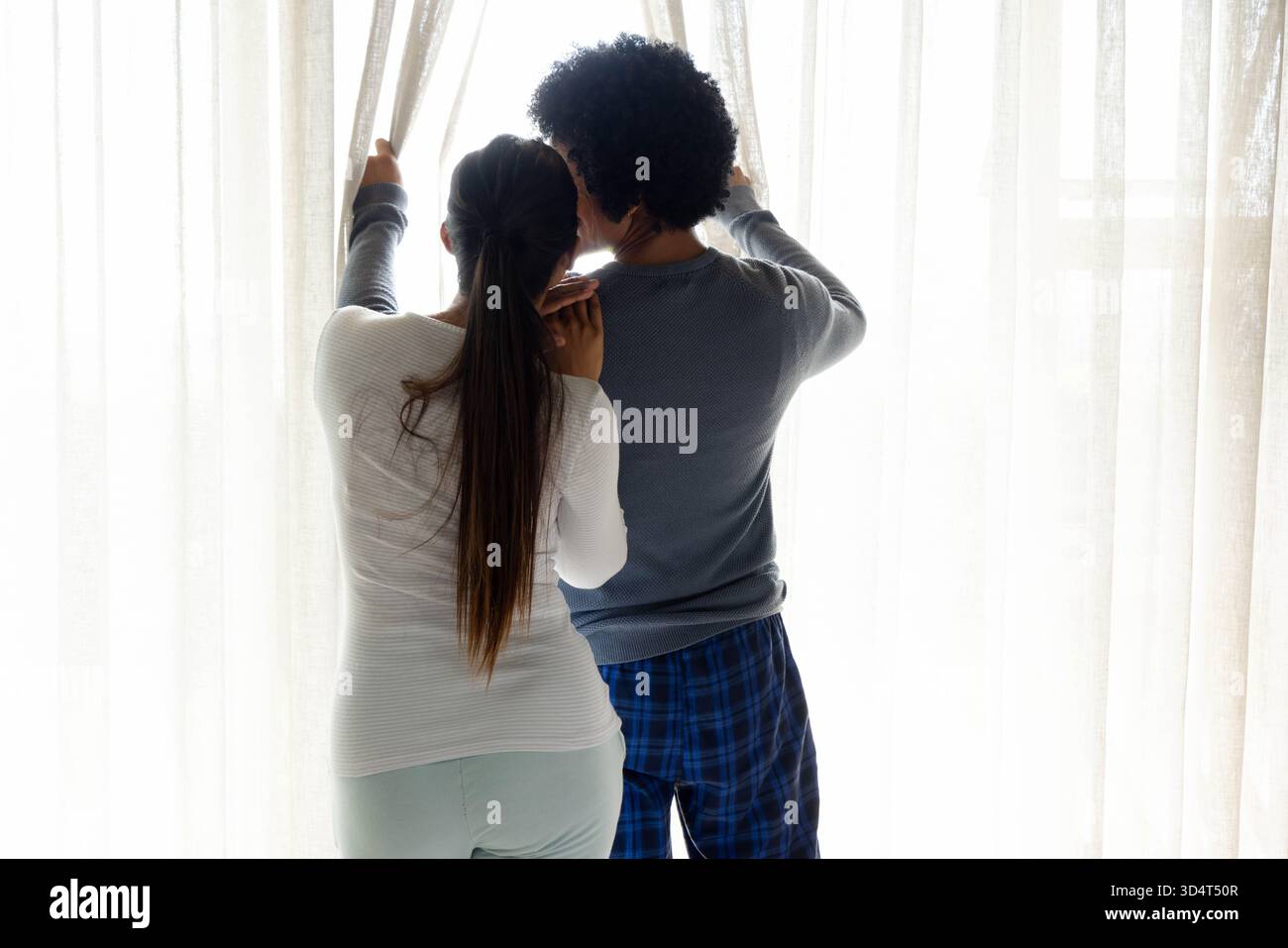 Tirando da parte le tende trasparenti diverse coppie in piedi davanti a una finestra a casa con lo streaming della luce del giorno Foto Stock