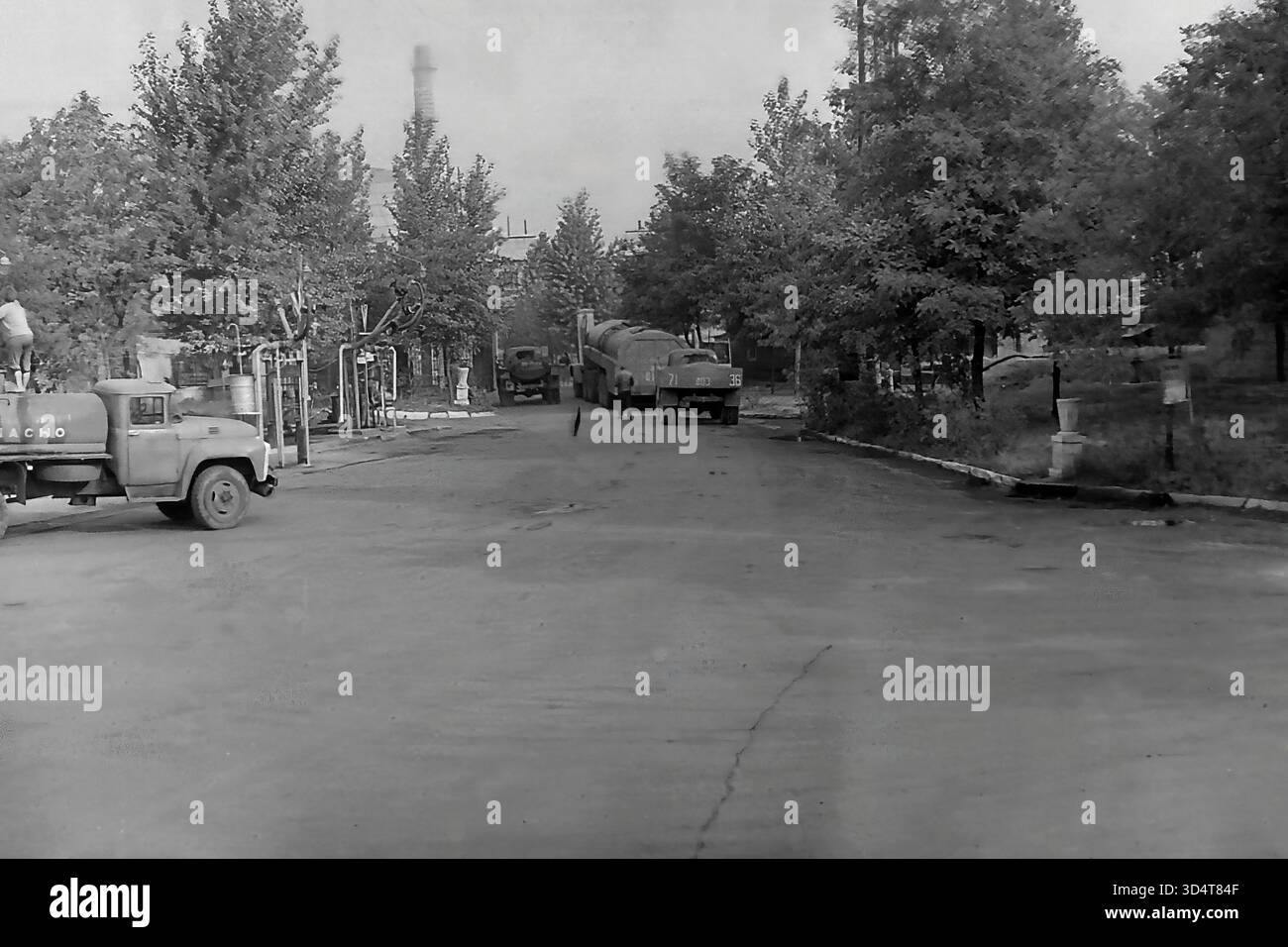 Una foto d'archivio in bianco e nero degli anni '1970 mostra una vista generale del deposito petrolifero di Sloviansk, la RSS Ucraina. Diversi autocisterne sovietici sono parcheggiati lungo una strada asfaltata costeggiata da alti alberi frondosi, con un lavoratore visibile su una cisterna. Sullo sfondo, un'alta cisterna industriale sorge sopra la struttura, definendo il paesaggio. Questa immagine cattura la logistica di routine e la miscela unica di industria e natura all'interno del pacifico Donbas durante l'era sovietica. Foto Stock