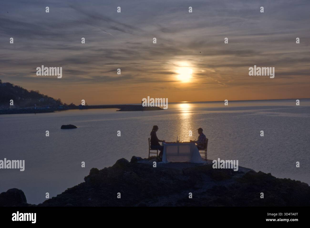 Una scena mozzafiato di una coppia che si gode una cena romantica privata su una scogliera rocciosa che si affaccia sul mare calmo al tramonto. Le cifre e la tabella set Foto Stock