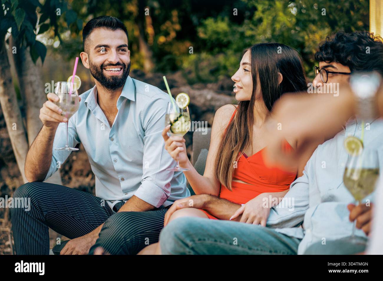 Un gruppo felice e multietnico di giovani amici siede all'aperto, parlando e bevendo cocktail a una festa in giardino estiva. Foto Stock