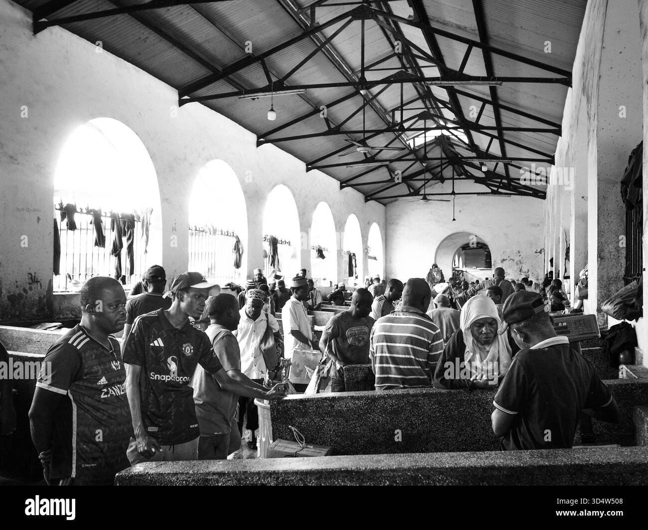 Sansibar, Tansania, Afrika: Innenansicht der Haupthalle des Darajani-Marktes Fischmarkt. Zahlreiche Personen sind in der Halle zwischen den niedrigen Verkaufstischen versammelt. Die Architektur ist durch hohe Wände mit bogenförmigen Öffnungen und ein offenes Dachgebälk gekennzeichnet, das helles Licht in den Raum lässt. Die Szene ist in Schwarz-Weiß gehalten. *** Zanzibar, Tanzania, Africa Vista interna della sala principale del mercato del pesce di Darajani numerose persone sono riunite nella sala tra i bassi tavoli di vendita l'architettura è caratterizzata da alte pareti con aperture ad arco e Foto Stock