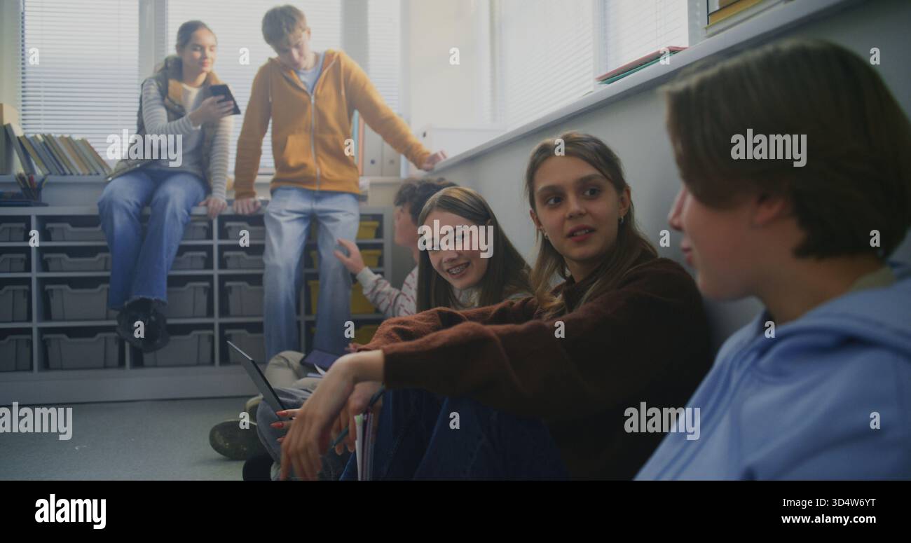 Gli studenti si siedono seduti sul pavimento attorno al laptop, condividendo idee e scrivendo note durante la sessione di studio collaborativo. Concetto di apprendimento tra pari, dialogo aperto e ambiente moderno e flessibile in aula. Foto Stock