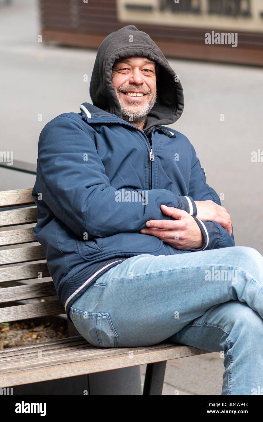 Un uomo di 50-55 anni con giacca e cappuccio blu è seduto su una panchina in città Foto Stock