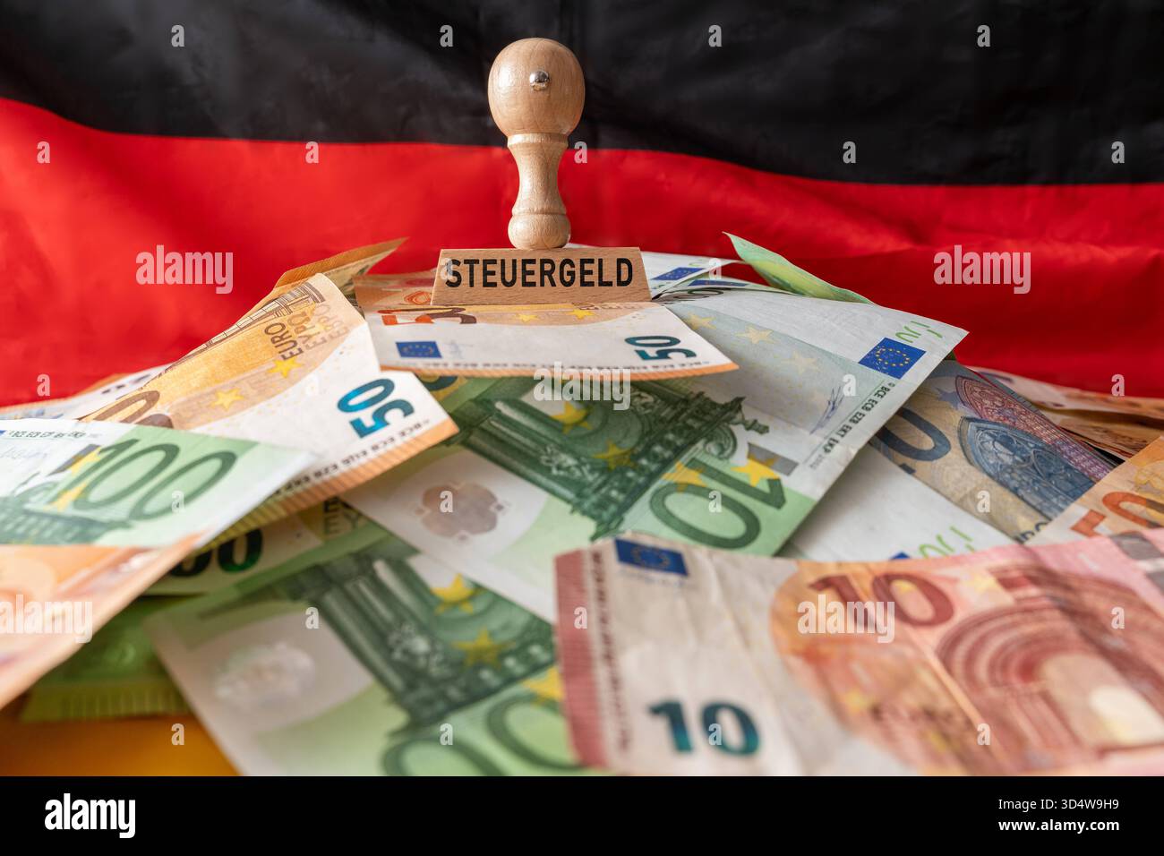 Baviera, Germania - 10 novembre 2025: MONTAGGIO FOTOGRAFICO, concetto relativo al gettito fiscale e all'utilizzo dei fondi pubblici: Un francobollo recante la dicitura gettito fiscale su una pila di banconote in euro davanti a una bandiera tedesca rappresenta le entrate, le spese e il dibattito sull'uso efficiente delle entrate fiscali. *** FOTOMONTAGE, Konzept zu Steuergeld und Verwendung öffentlicher Mittel: Ein Stempel mit der Aufschrift Steuergeld auf einem Haufen Euro-Banknoten vor einer Deutschlandflagge steht für staatliche Einnahmen, Ausgaben und die Debatte über effiziente Nutzung von Steuerteln. Foto Stock