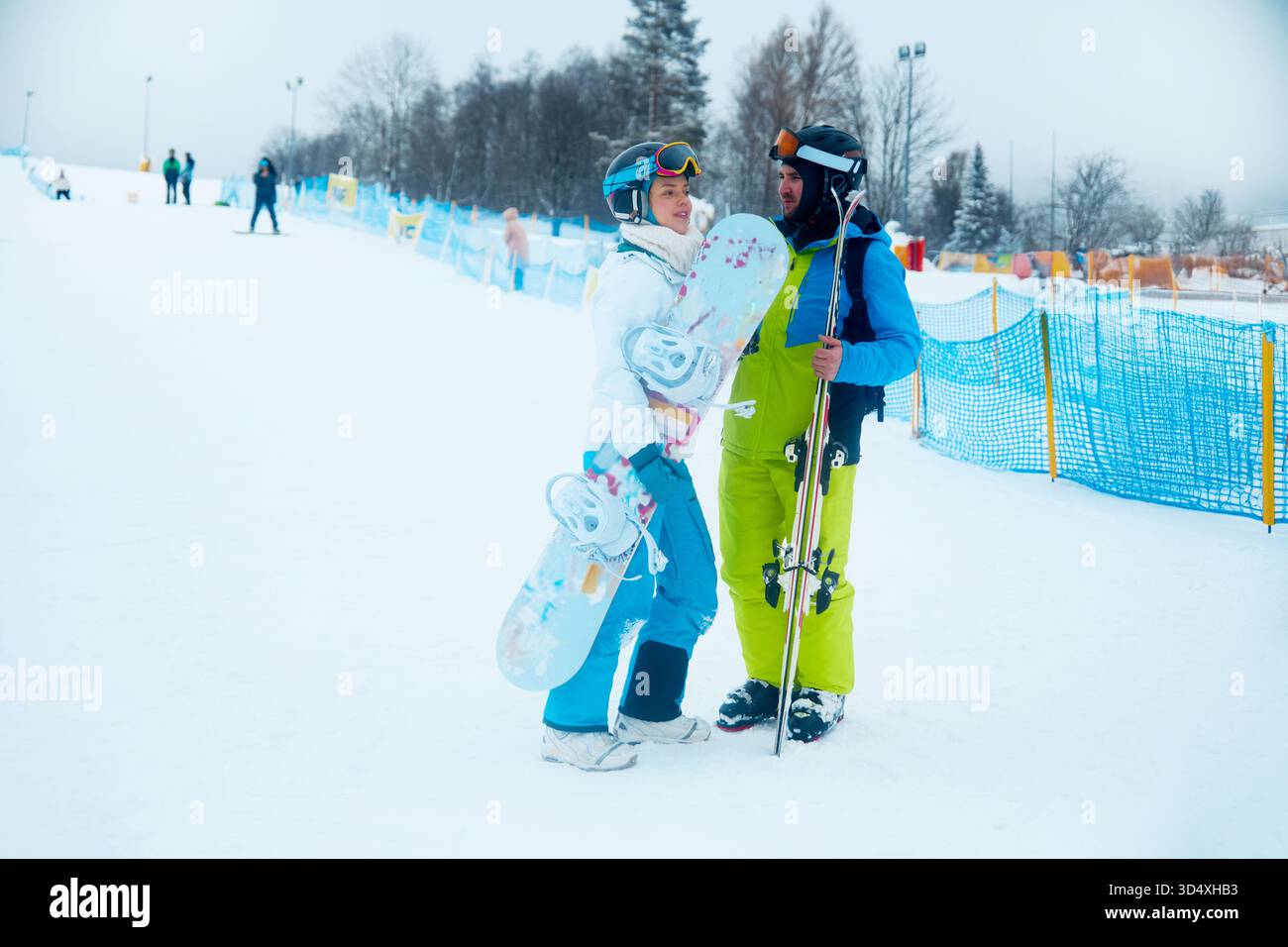Due amici condividono un momento di gioia in una stazione sciistica, tenendo snowboard e sci mentre sono circondati dalla neve e da altri sciatori in una giornata fredda. Foto Stock