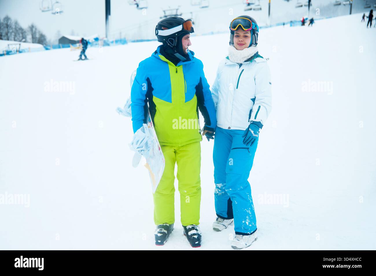 Una coppia si erge su una pista da sci innevata, vestita con abiti da sci colorati. Sorridono e si tengono per mano, emozionati per la loro avventura invernale. Foto Stock