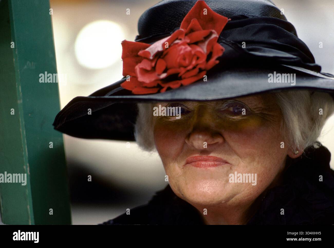 Donna anziana matura e sicura di sé che guarda la fotocamera. Elegante cappotto e cappello nero. Un grande fiocco rosso "rosa" attaccato al suo cappello a teglia larga. Signora con i capelli bianchi è auto-posseduta, Londra Inghilterra Regno Unito HOMER SYKES Foto Stock