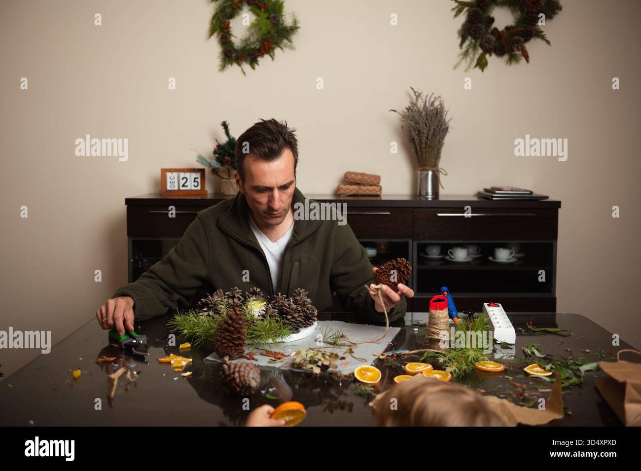 Un uomo crea decorazioni pinecone su un accogliente tavolo da pranzo, preparando ghirlande e accenti di vacanza in un ambiente caldo e familiare. Foto Stock