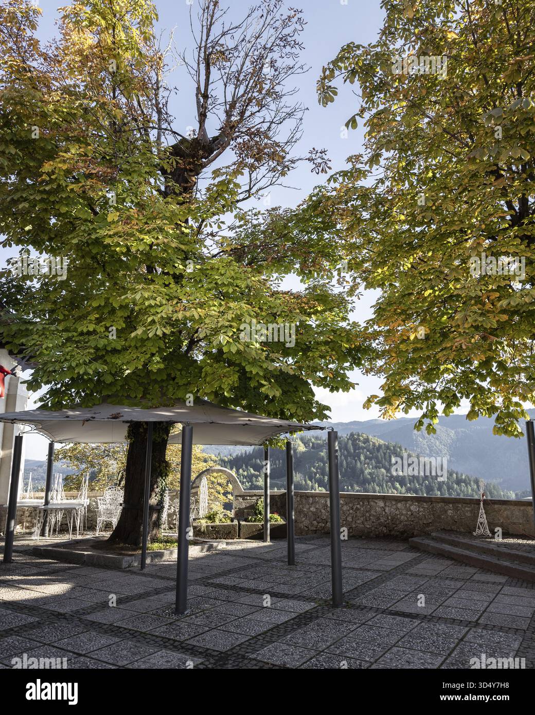 Vista delle foglie autunnali tra gli alberi che si affacciano sulle colline da una piazza di pietra con strutture metalliche, Bled, Radovljica, Slovenia. Foto Stock