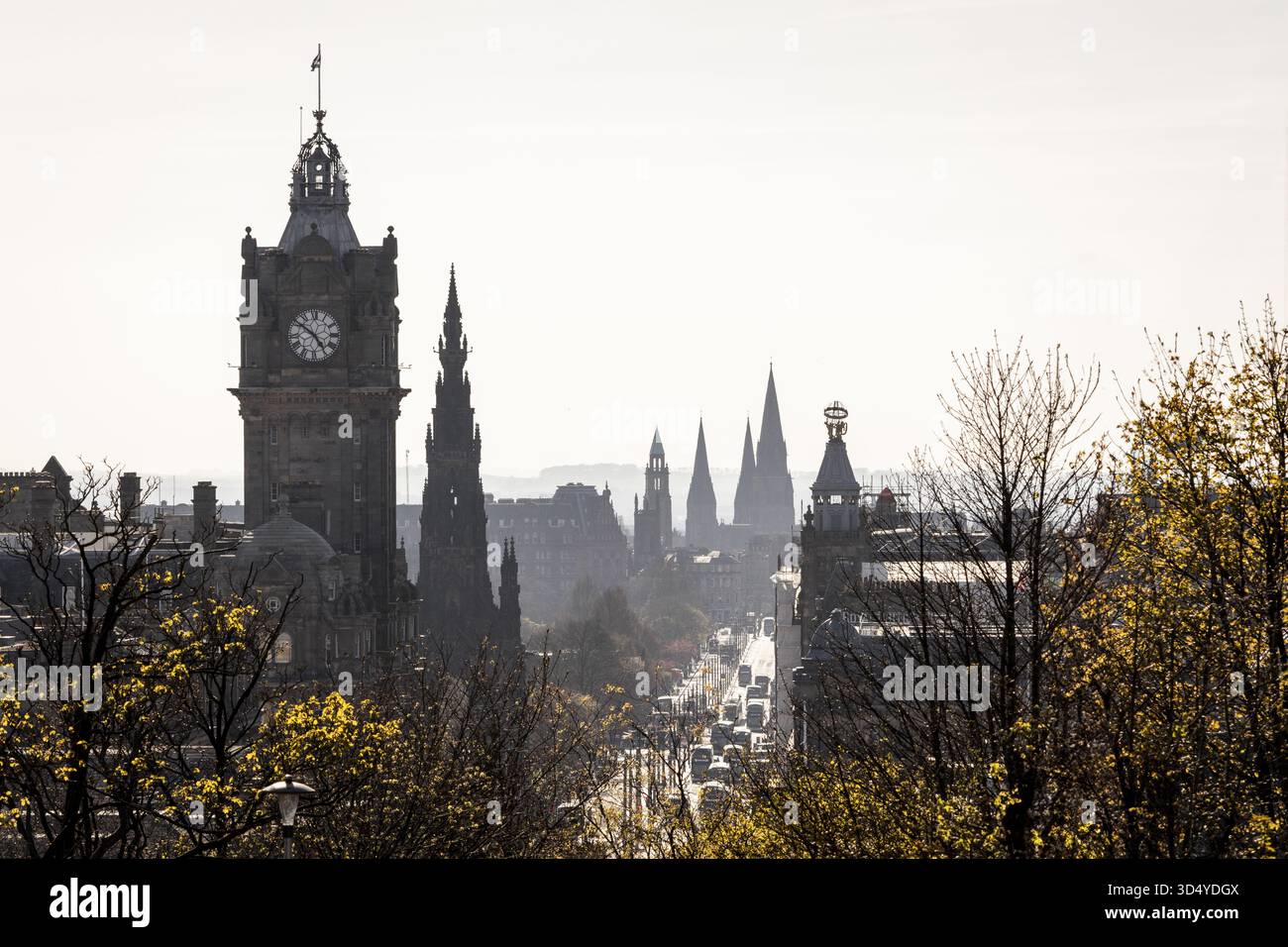 La vista dell'iconica torre dell'orologio Balmoral e delle guglie gotiche perforano lo skyline nebbioso, incorniciato da alberi in erba lungo una vivace strada, Edimburgo, Scotla Foto Stock